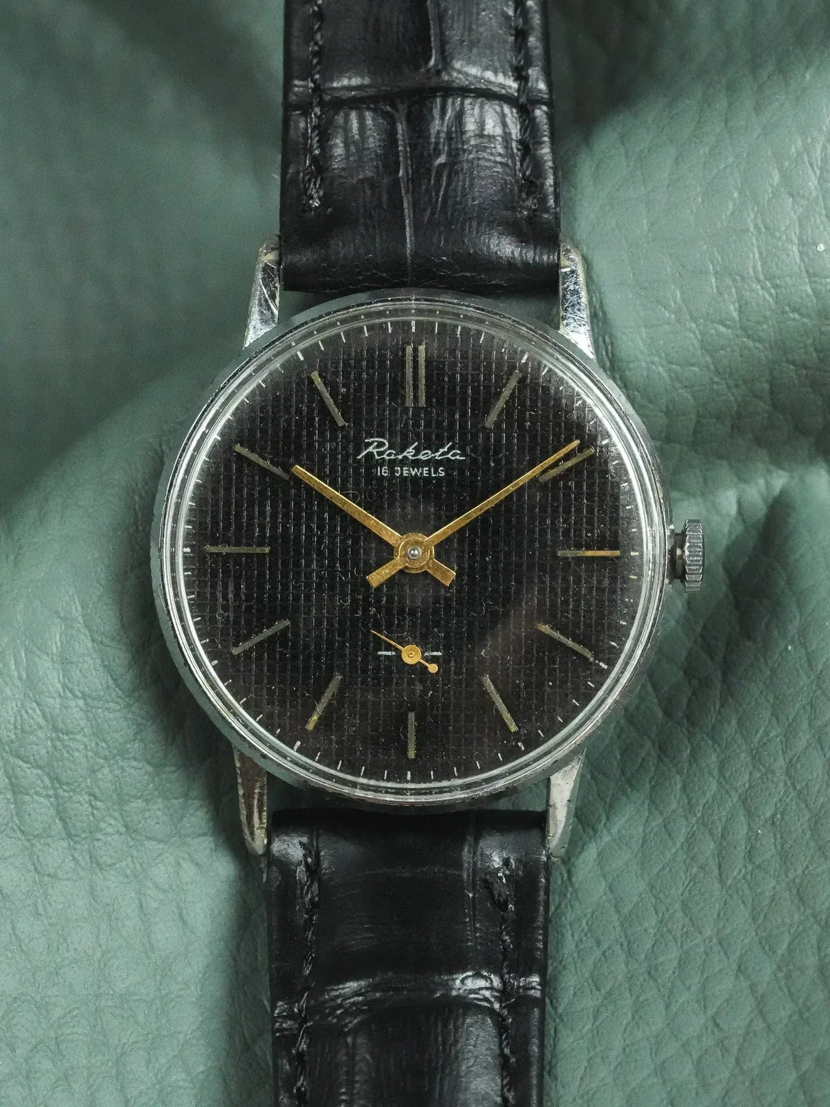 Raketa vintage watch with black textured dial, gold hands, and black leather strap
