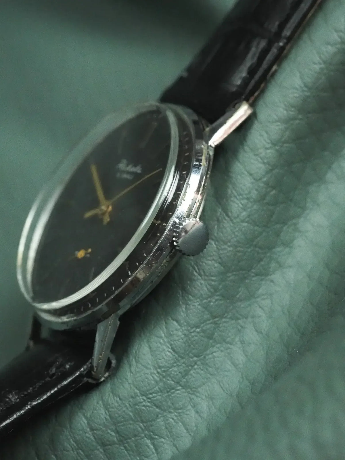 Vintage Rolex watch with black dial, gold hands, and black leather strap on green leather
