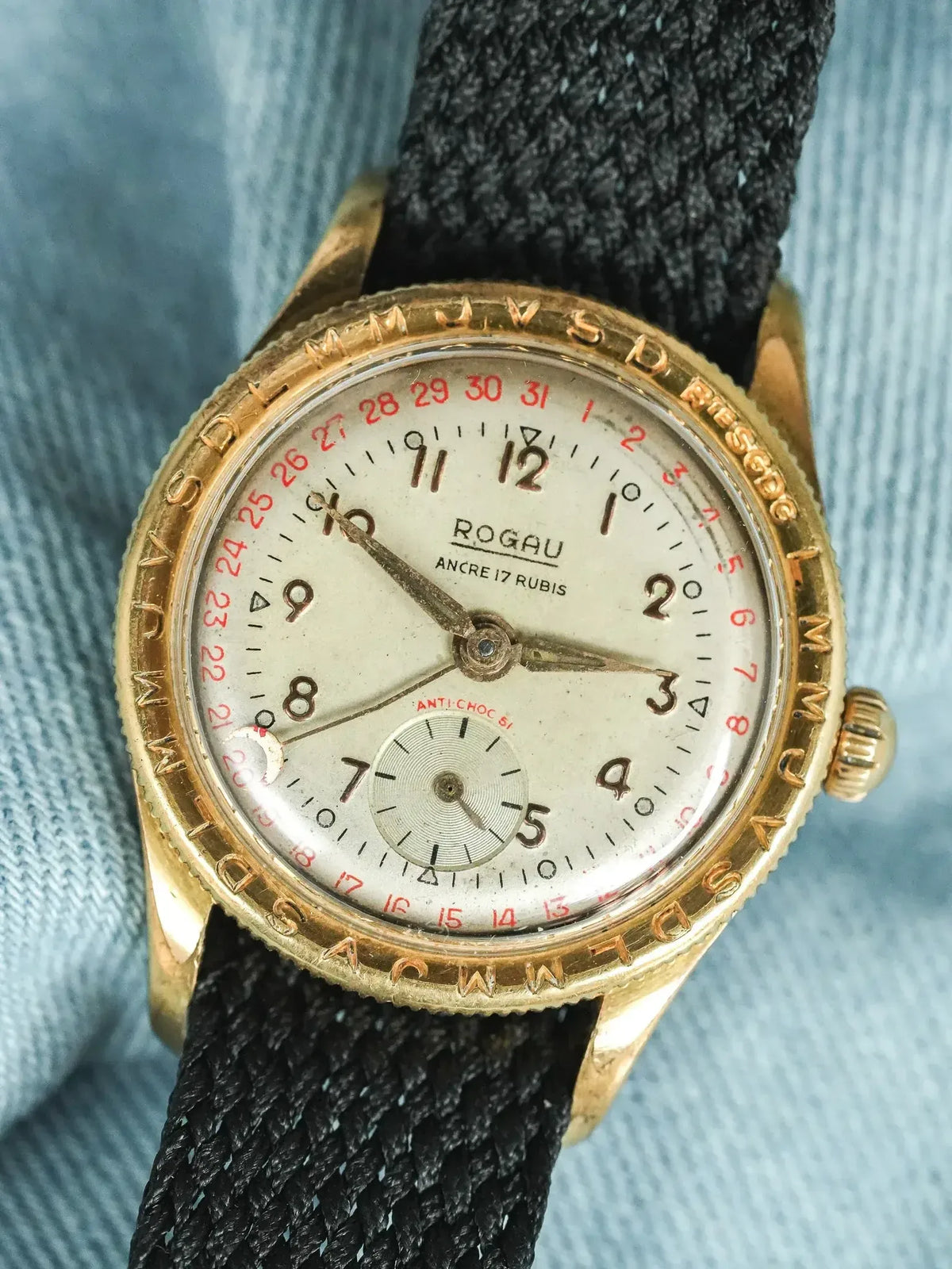 Vintage Rogau wristwatch with gold case, cream dial, red date markers, black fabric strap