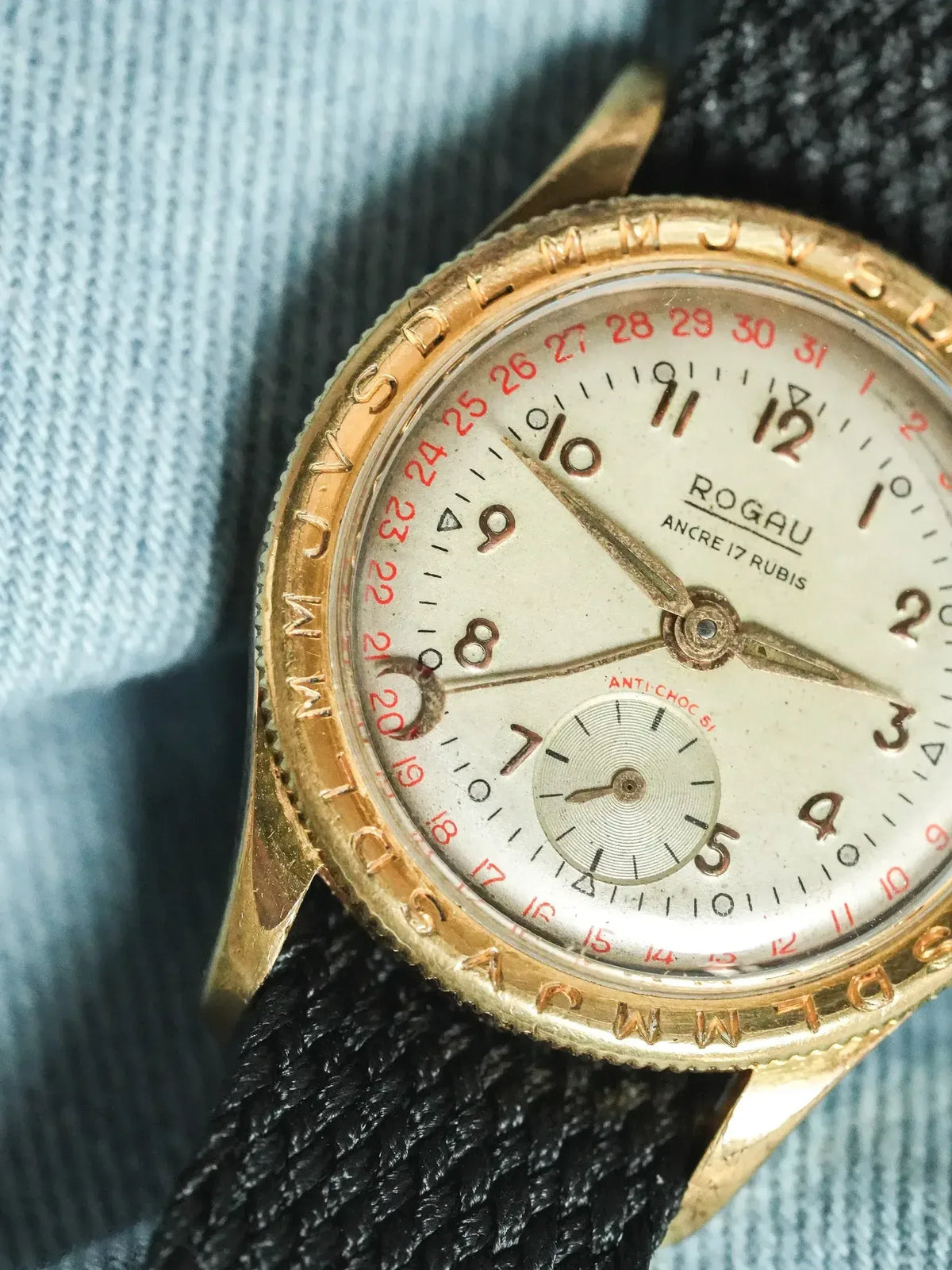 Rogau vintage watch with gold case, white dial, red date, black strap on denim