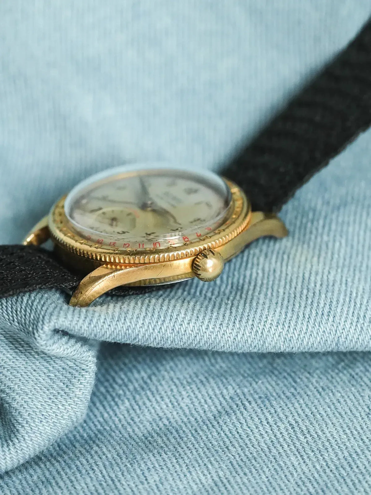 Vintage Rolex watch with gold case, cream dial, black fabric strap on denim