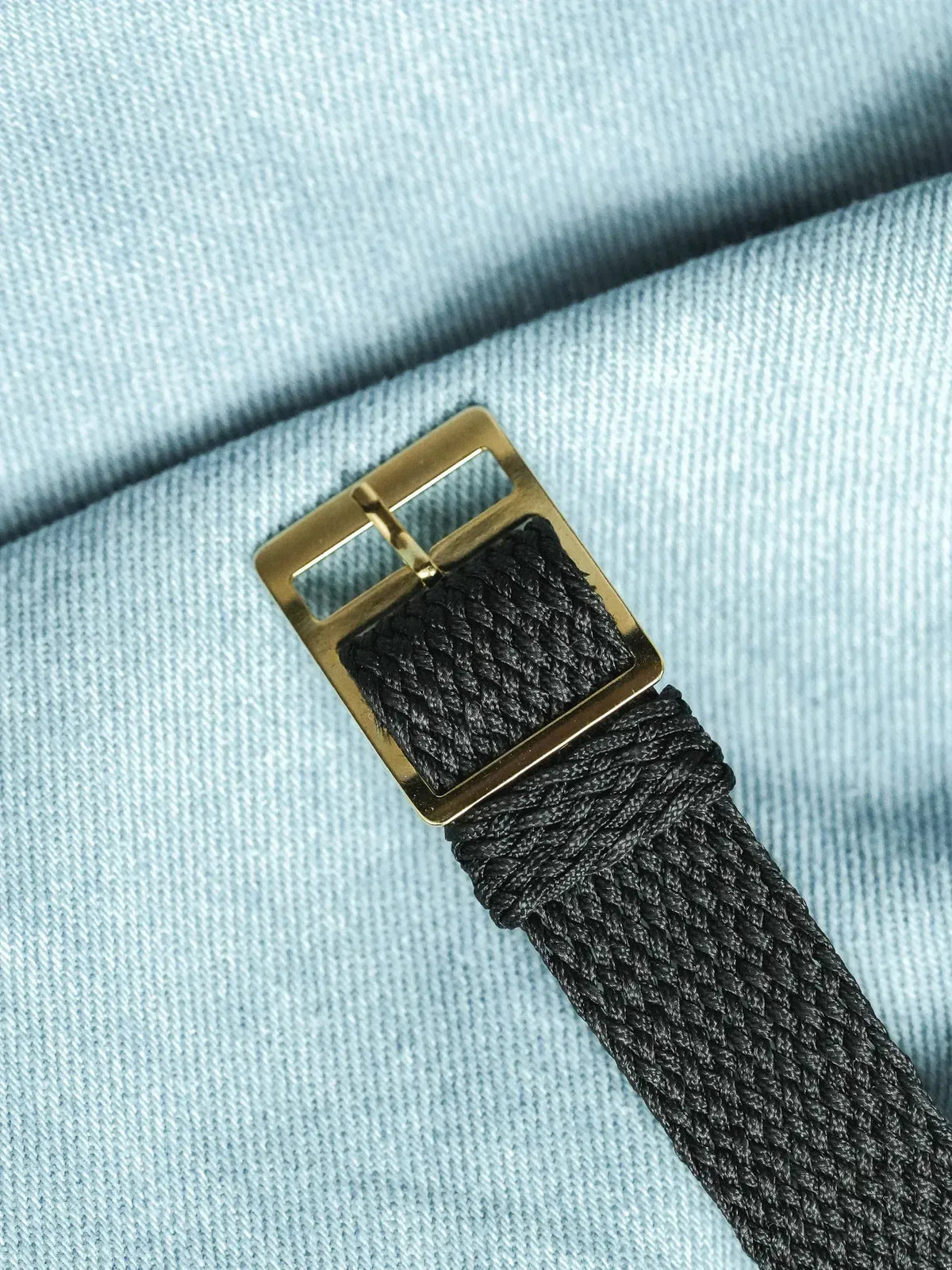 Black braided watch strap with gold buckle on light blue fabric