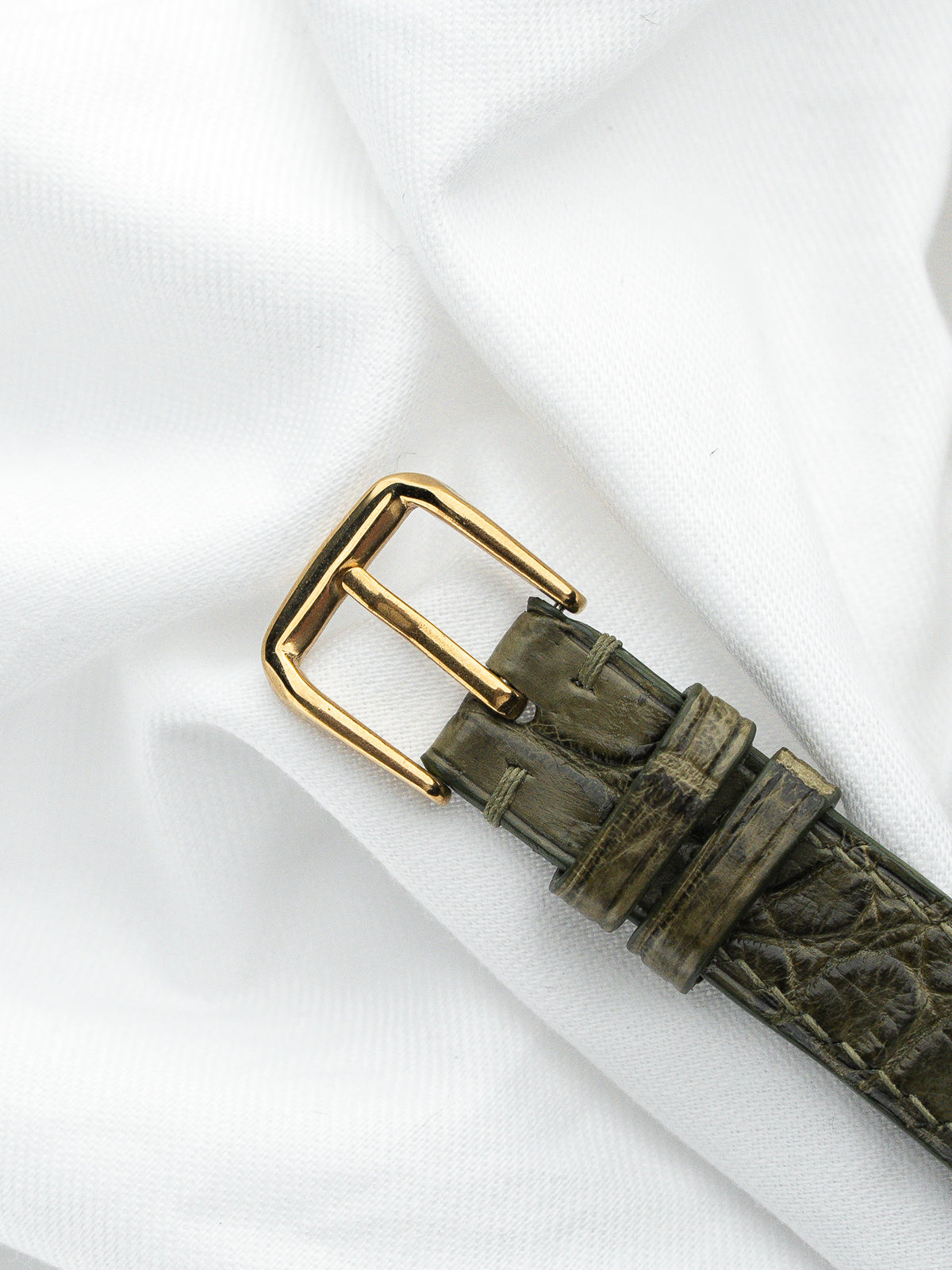 Détail d'un bracelet de montre en cuir vert olive avec boucle dorée sur tissu blanc.