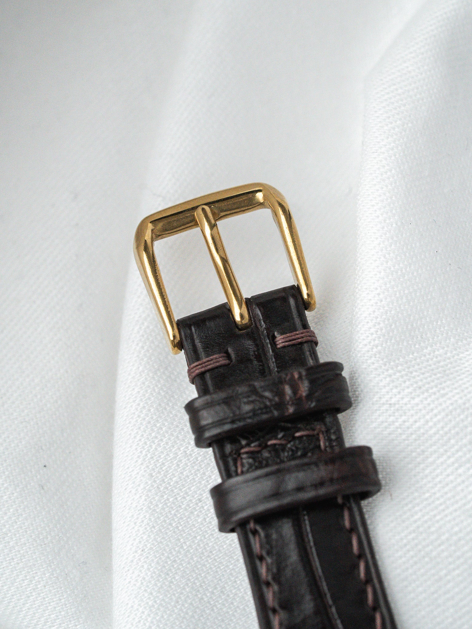 Vintage leather watch strap with gold buckle and brown stitching on white fabric
