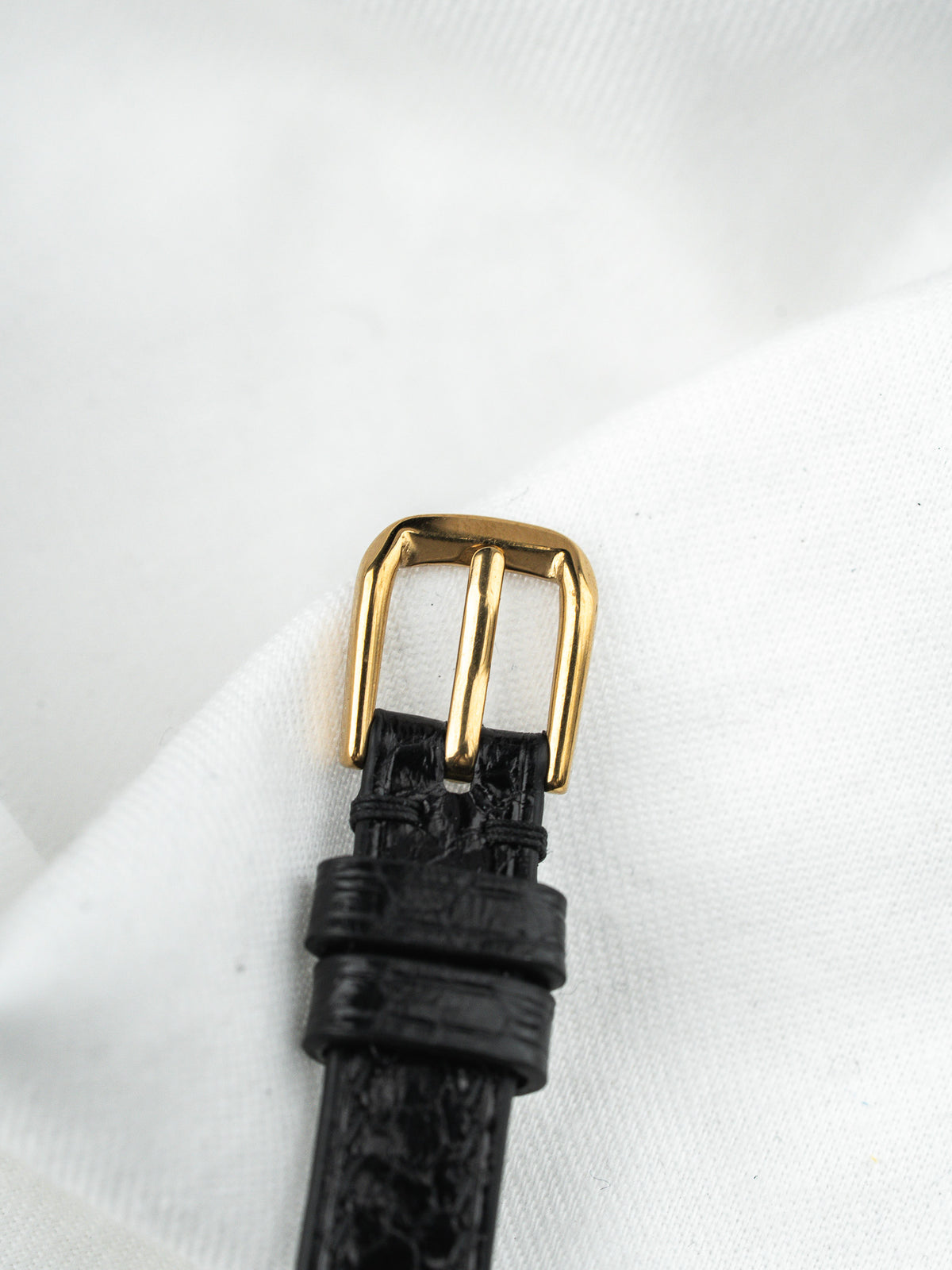 Boucle de bracelet de montre dorée sur cuir noir grainé, posée sur tissu blanc