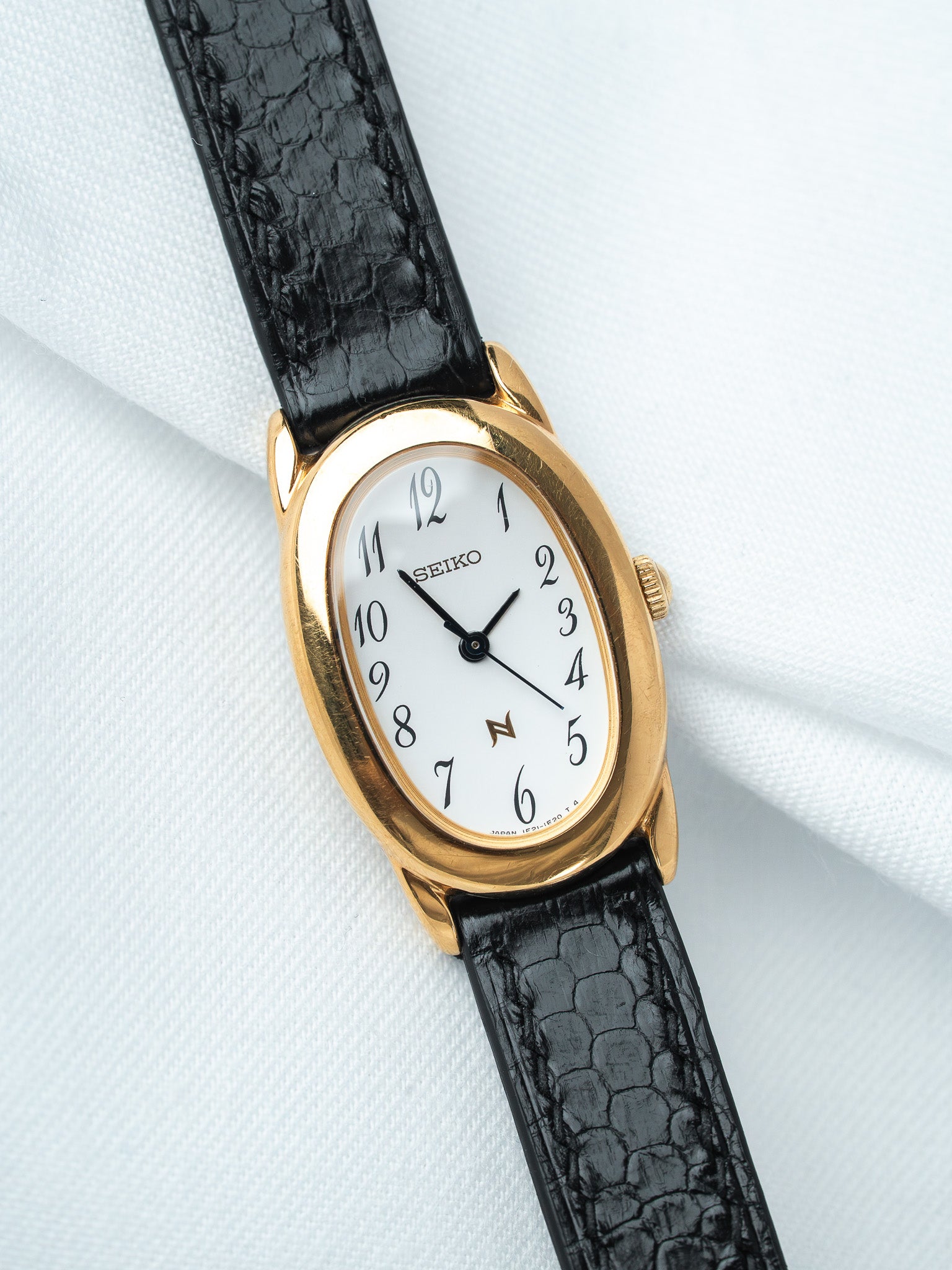 Seiko vintage watch with gold oval case, white dial, black crocodile strap on white fabric