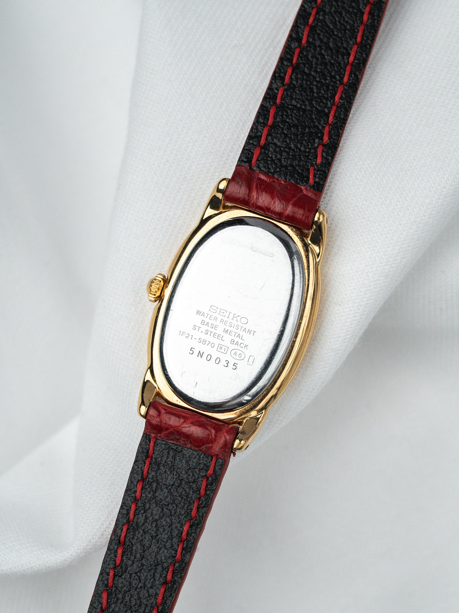 Seiko vintage watch with gold case, black leather strap, red stitching, displayed on white fabric