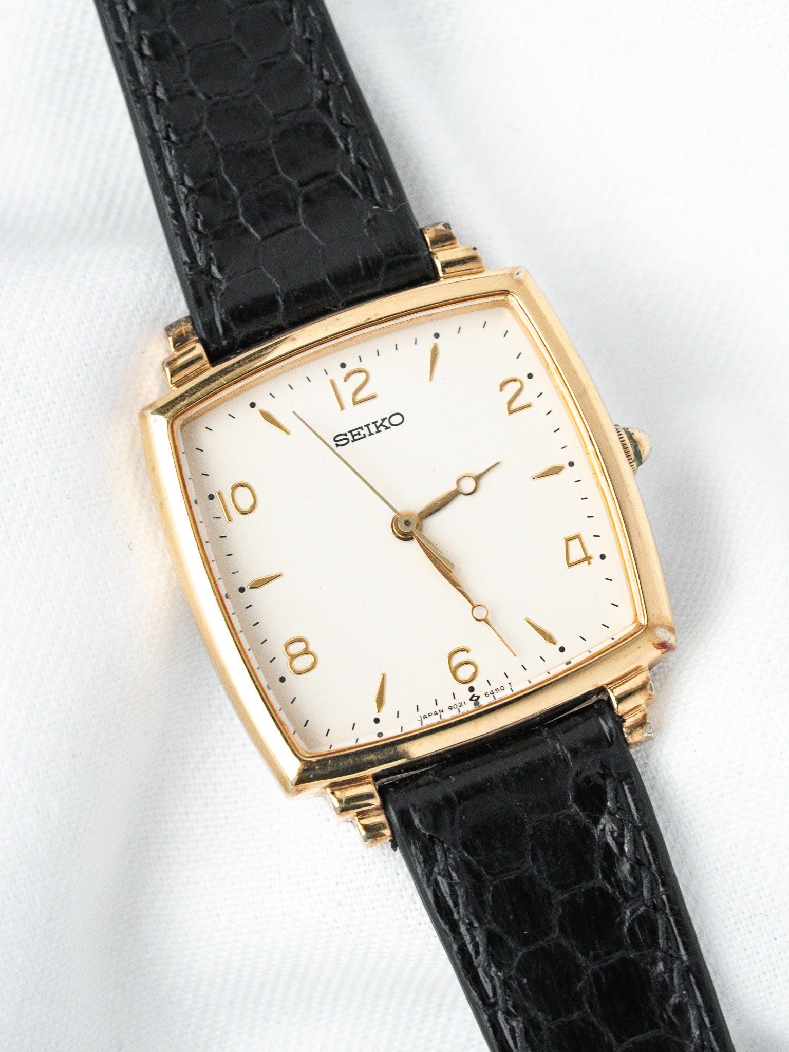 Seiko vintage watch with gold case, white dial, black alligator strap on white background