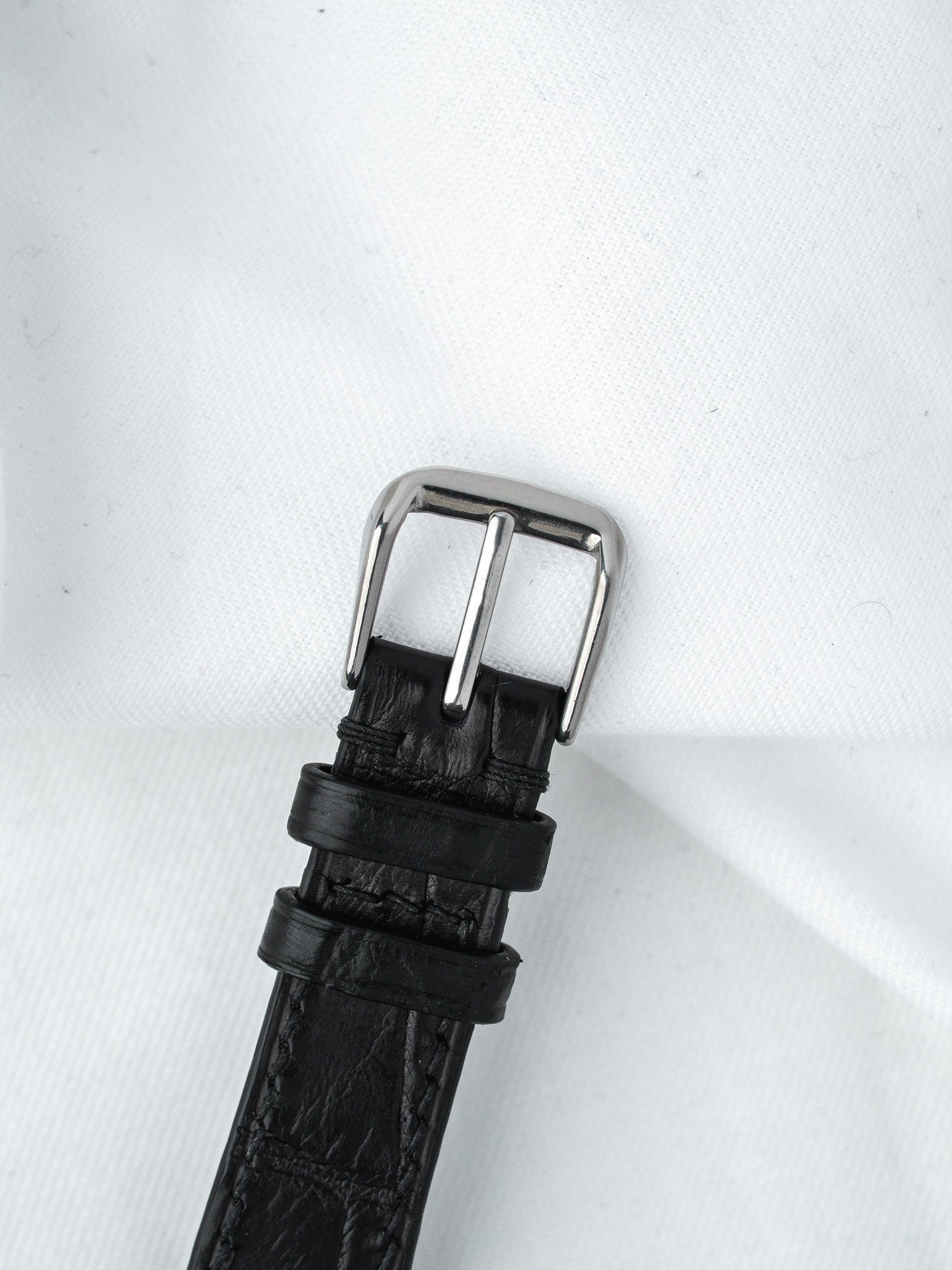 Black leather watch strap with silver buckle on white fabric background