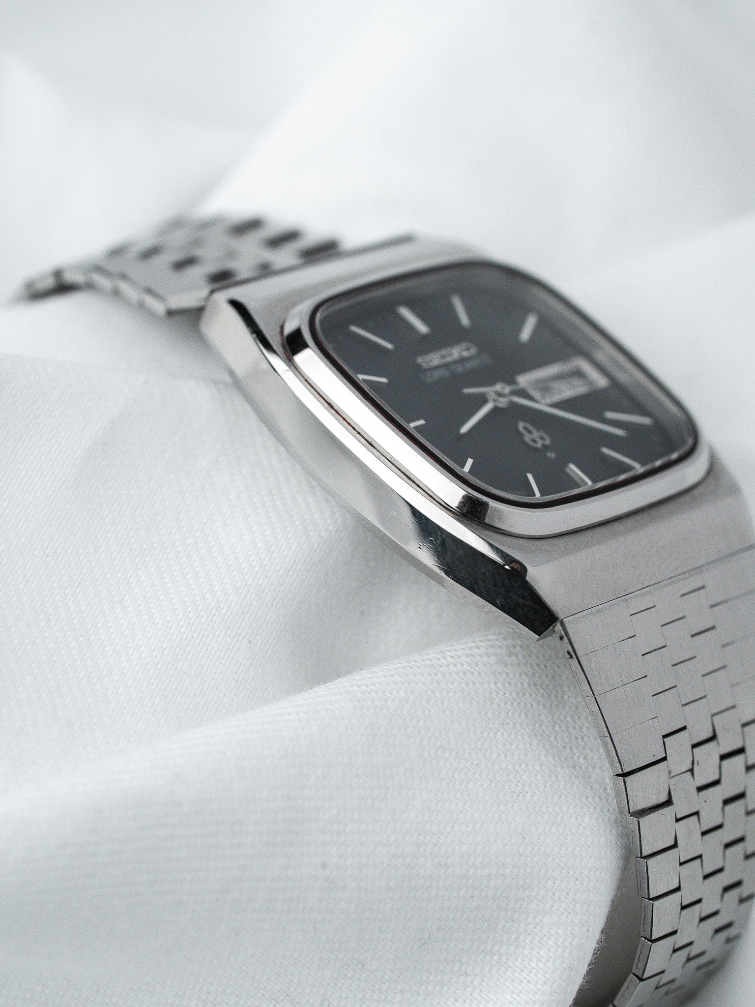 Seiko vintage watch with black dial, silver case, and woven bracelet on white fabric