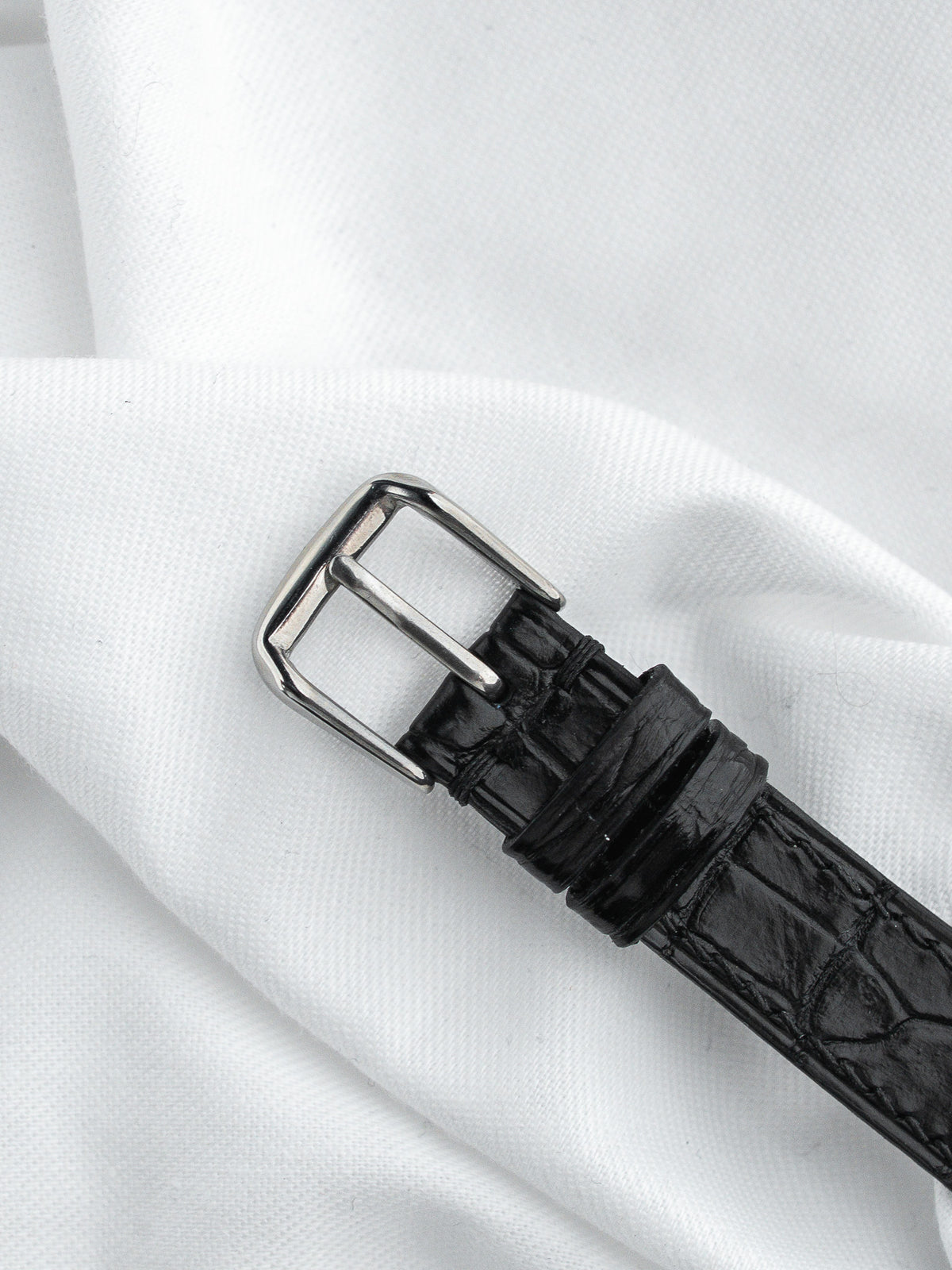 Boucle ardillon en acier et bracelet en cuir noir effet croco sur tissu blanc.