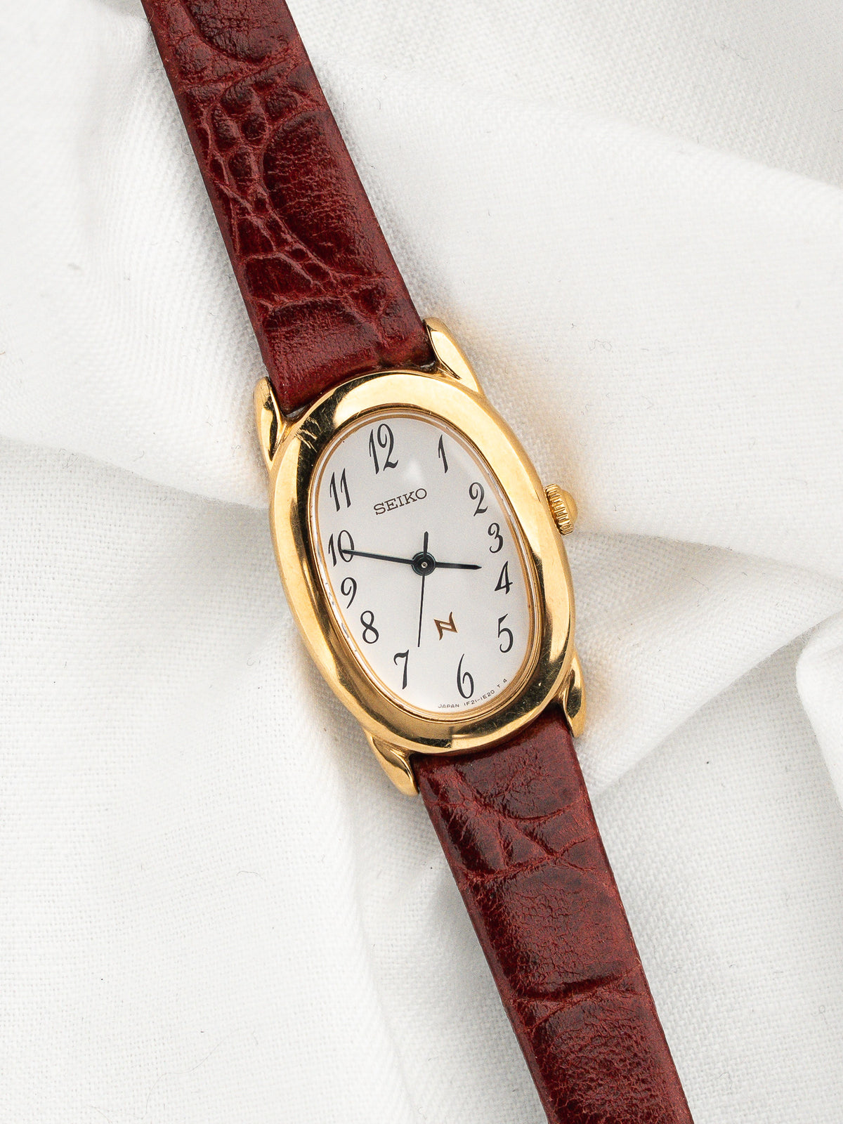 Montre Seiko vintage avec cadran blanc, boîtier doré et bracelet en cuir bordeaux.