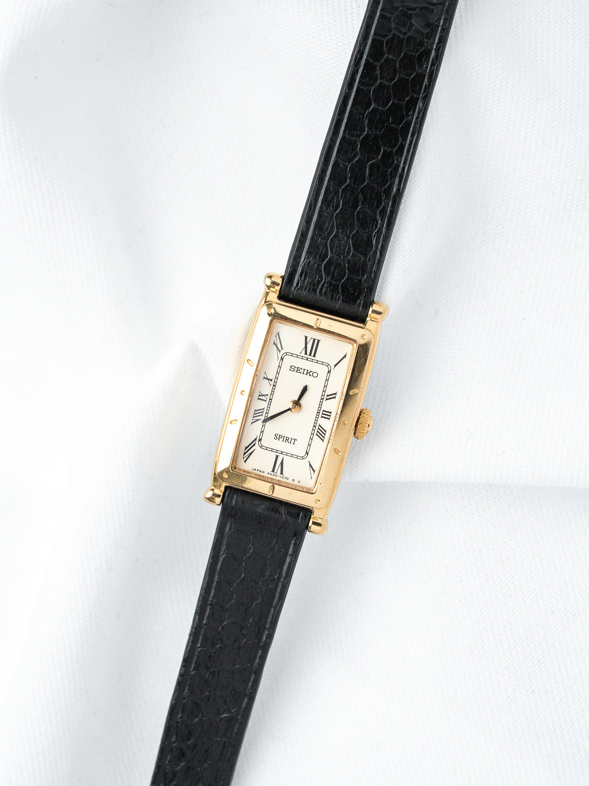 Seiko Spirit vintage watch with gold case, white dial, Roman numerals, black textured strap on white background