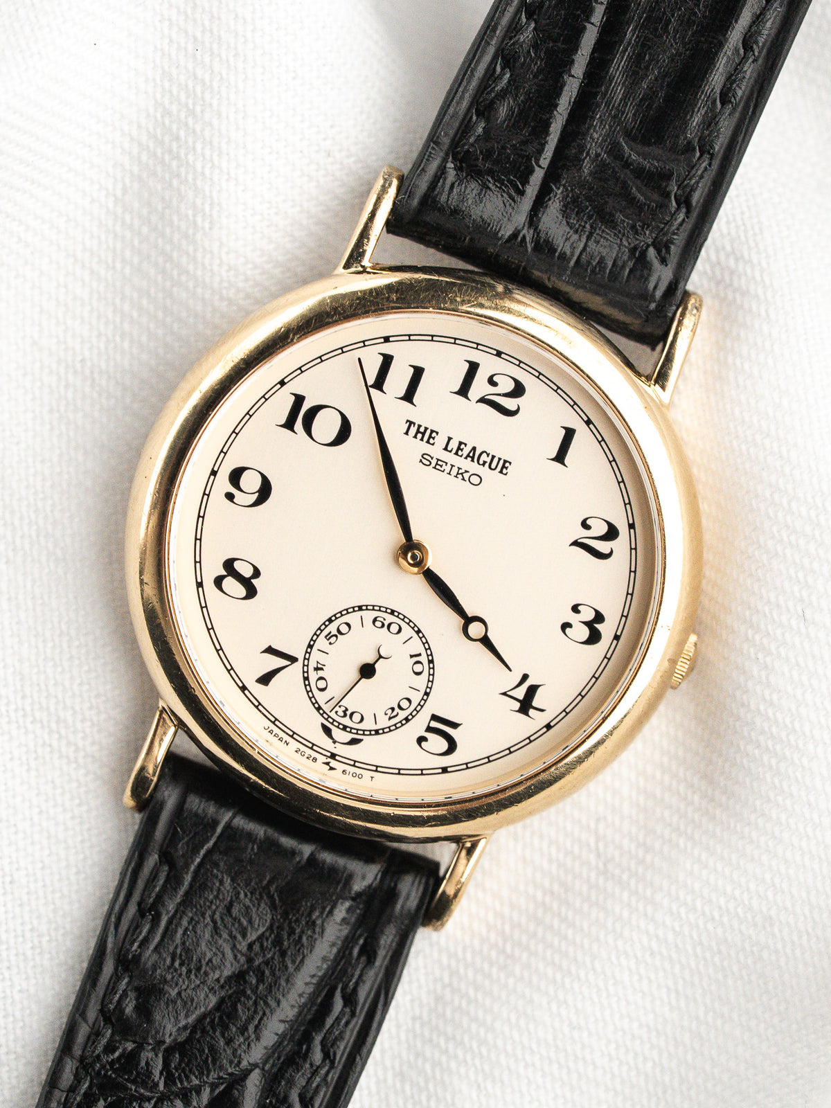 Montre Seiko The League avec cadran crème, boîtier doré et bracelet en cuir noir.