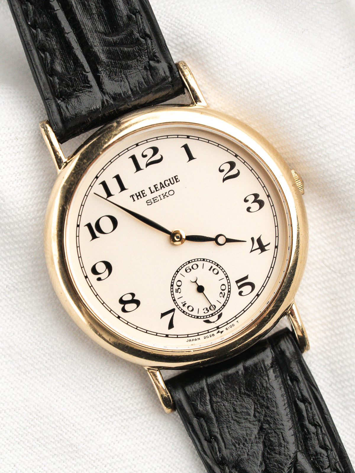 Montre Seiko The League vintage avec boîtier doré, cadran blanc et bracelet noir.