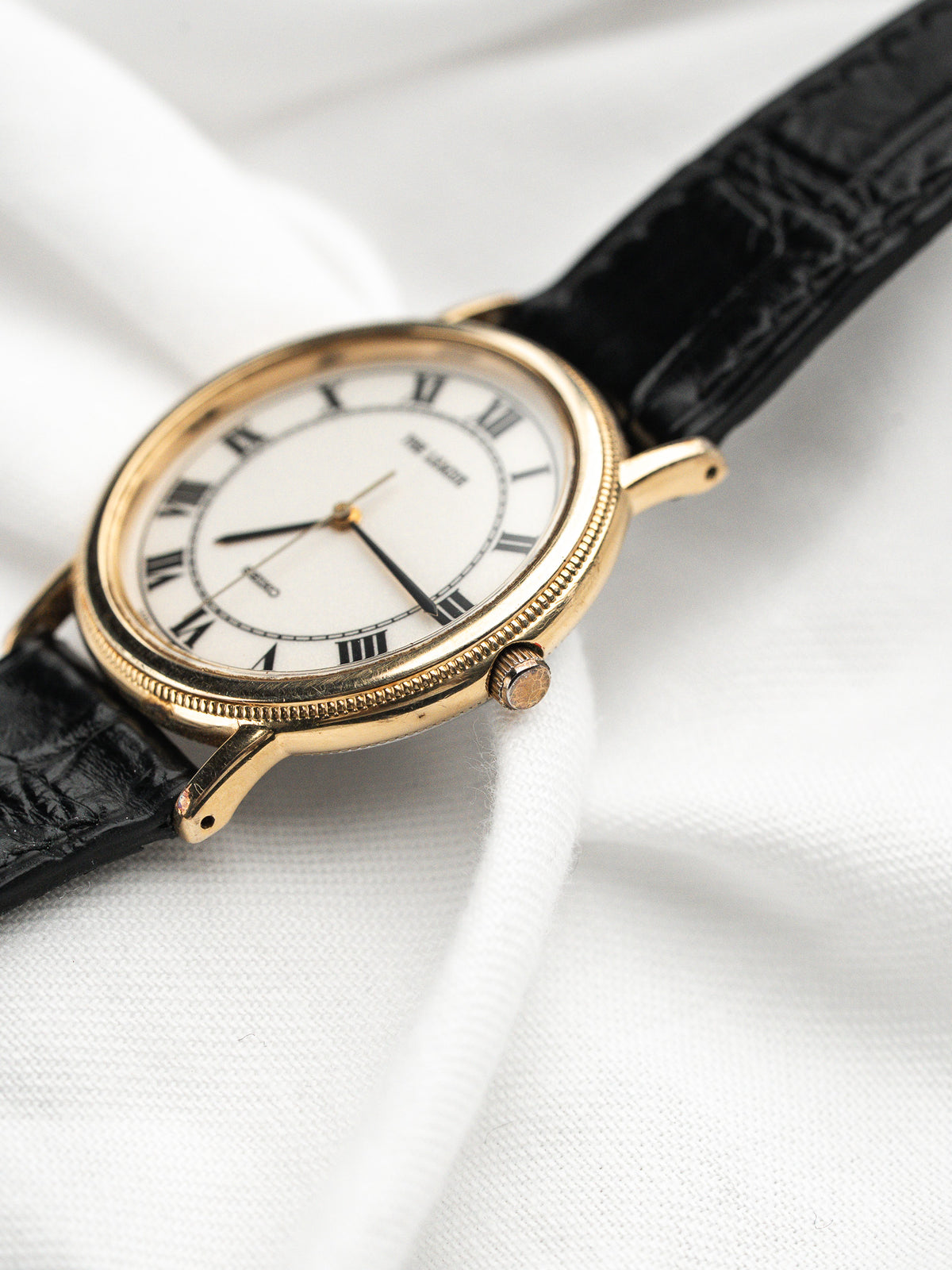 Montre vintage au boîtier doré, cadran blanc avec chiffres romains et bracelet cuir noir.