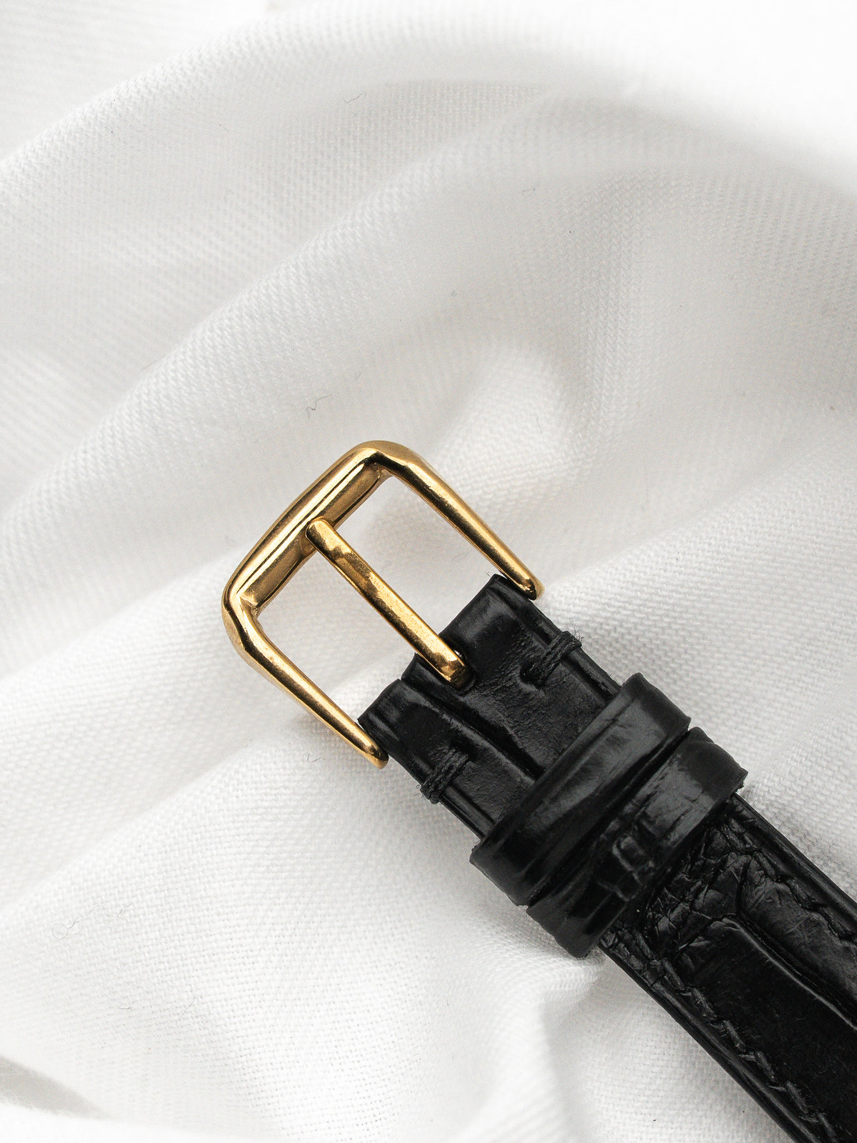 Boucle de montre dorée sur un bracelet en cuir noir posé sur tissu blanc.