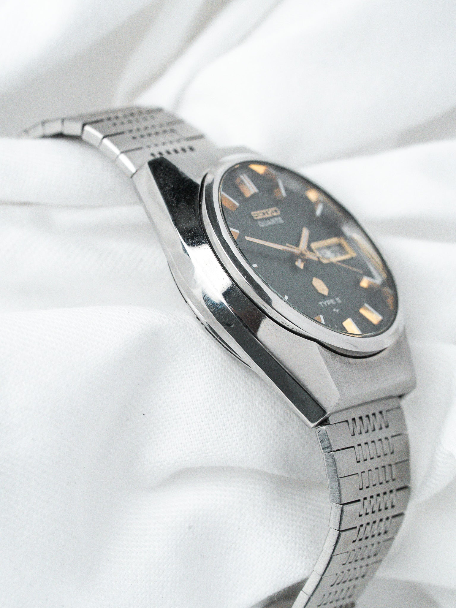 Seiko vintage quartz watch with black dial, gold markers, and steel bracelet on white fabric