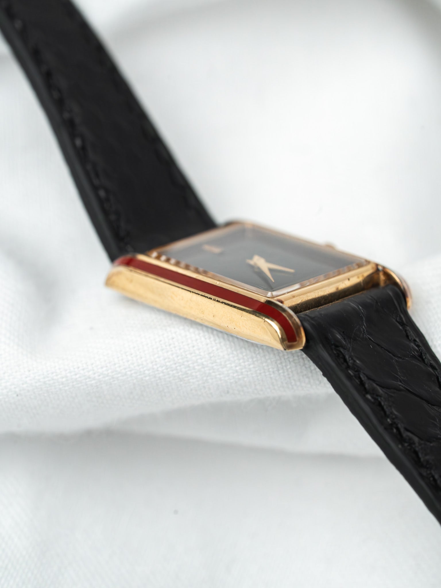 Vintage Cartier watch with gold case, red enamel accents, black leather strap on white fabric