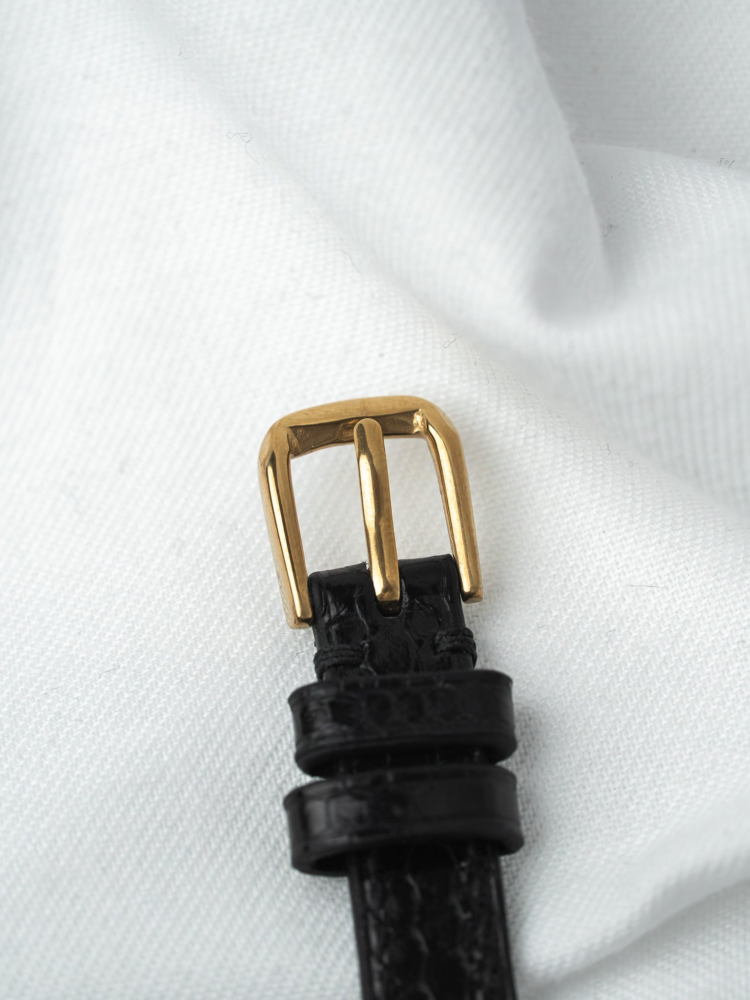 Black alligator strap with gold buckle on white fabric background