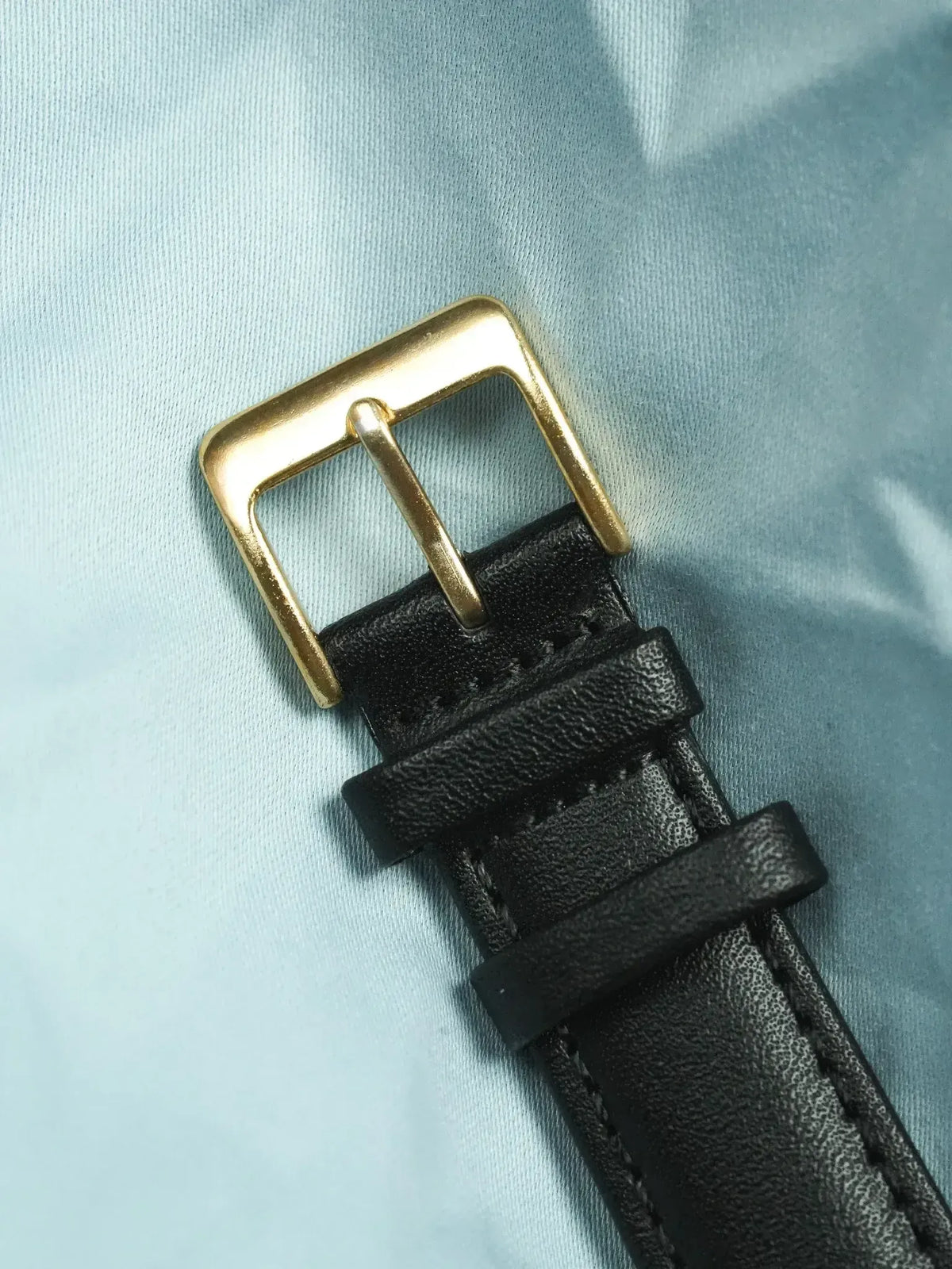 Black leather watch strap with gold-tone buckle on light blue fabric