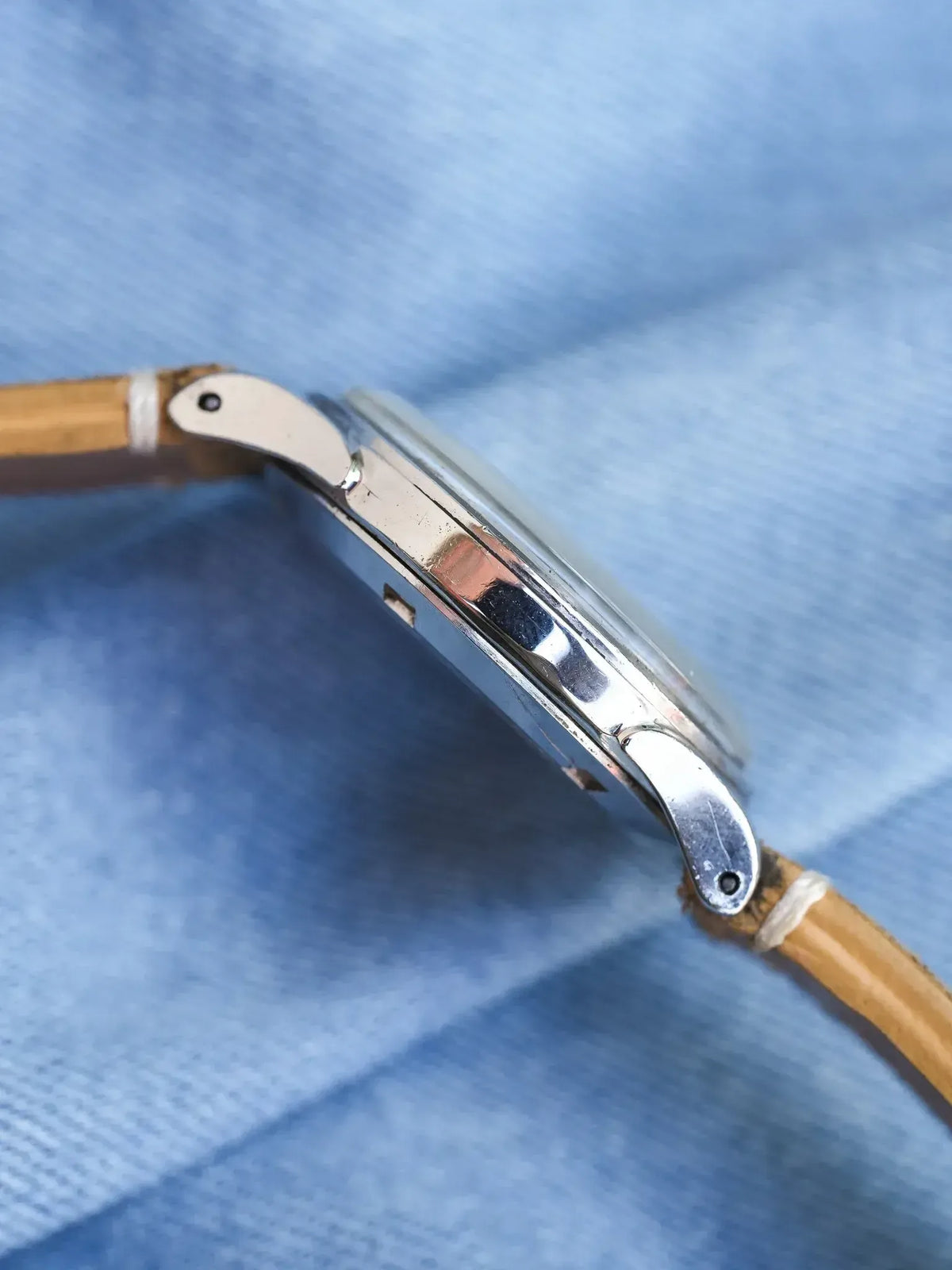 Vintage watch side profile with polished steel case and tan leather strap on blue fabric