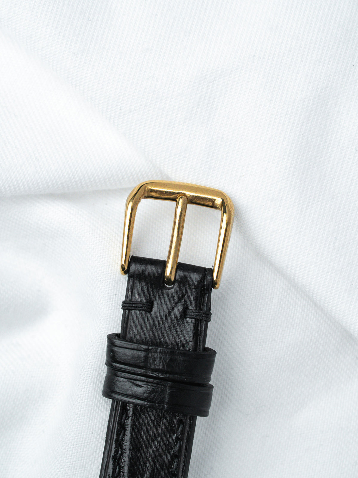 Black textured leather watch strap with gold buckle on white fabric