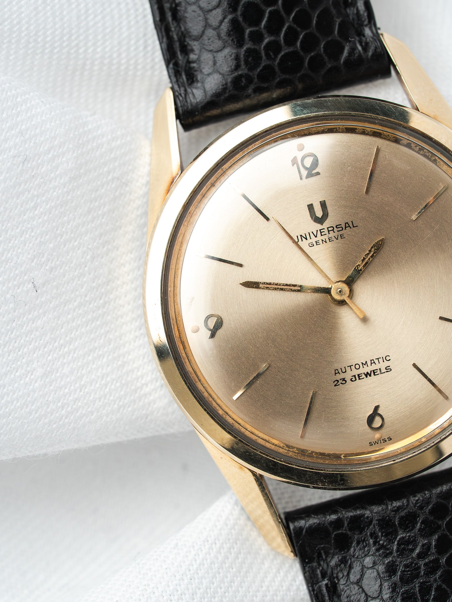 Universal Genève vintage watch with gold case, silver dial, black leather strap on white fabric
