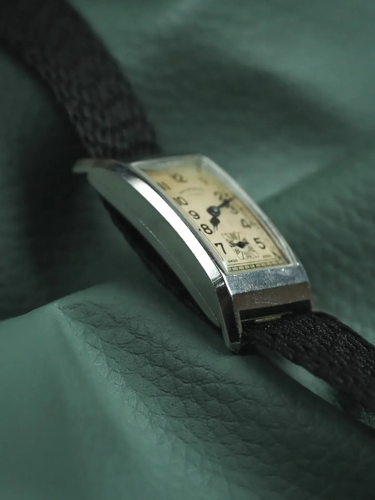 Vintage rectangular watch with cream dial, black numerals, and black fabric strap on green leather