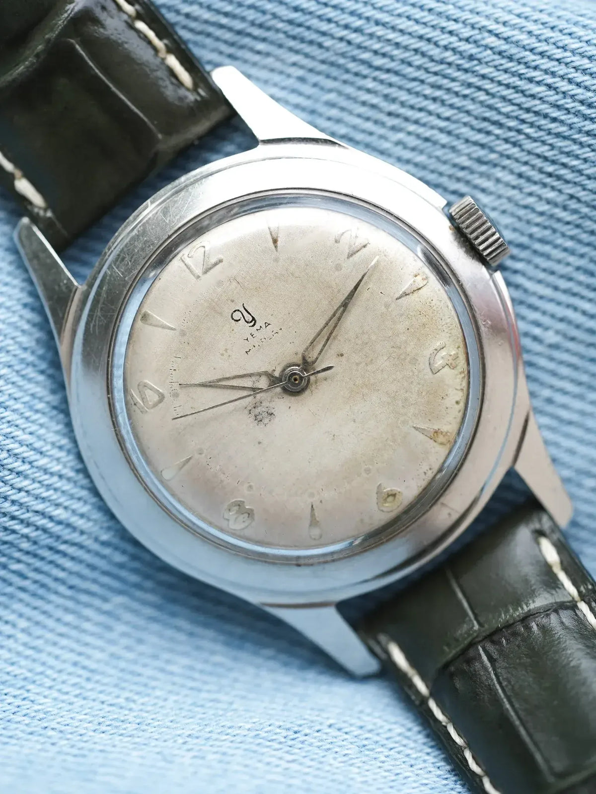 Vintage Yema watch with silver dial, black leather strap, resting on blue fabric