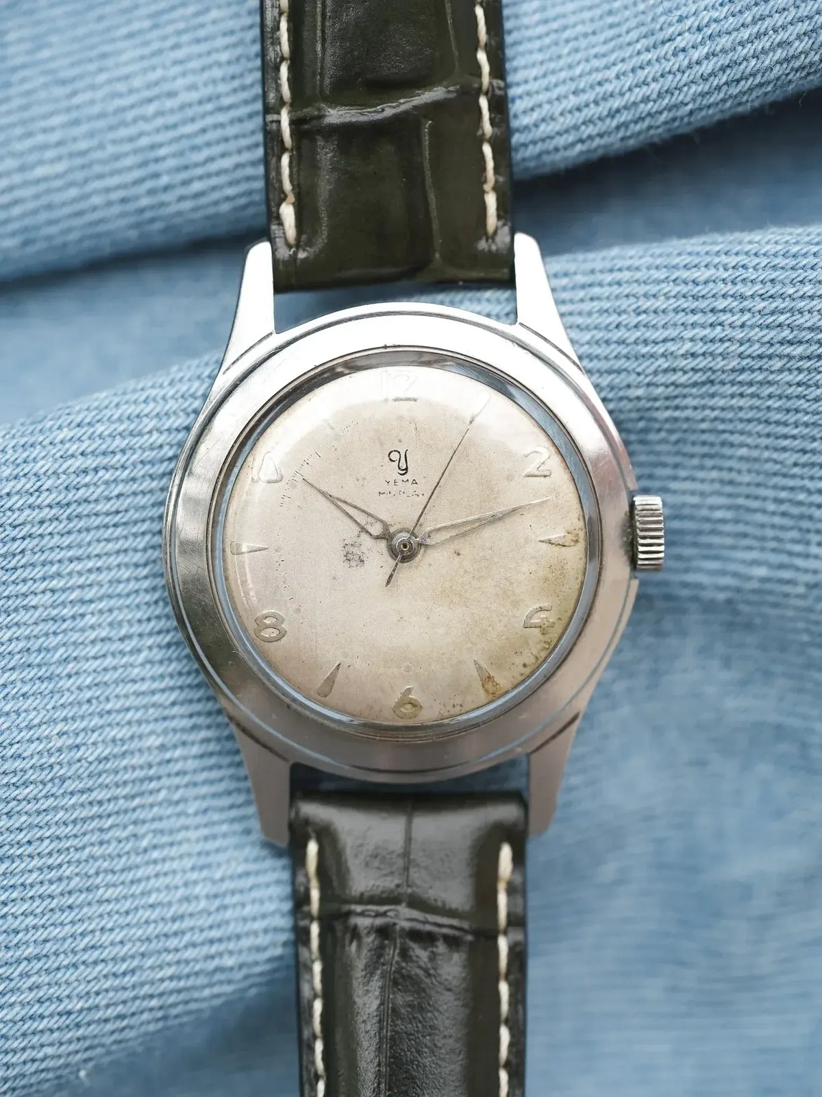 Vintage Yema watch with silvered dial, steel case, and dark leather strap on denim