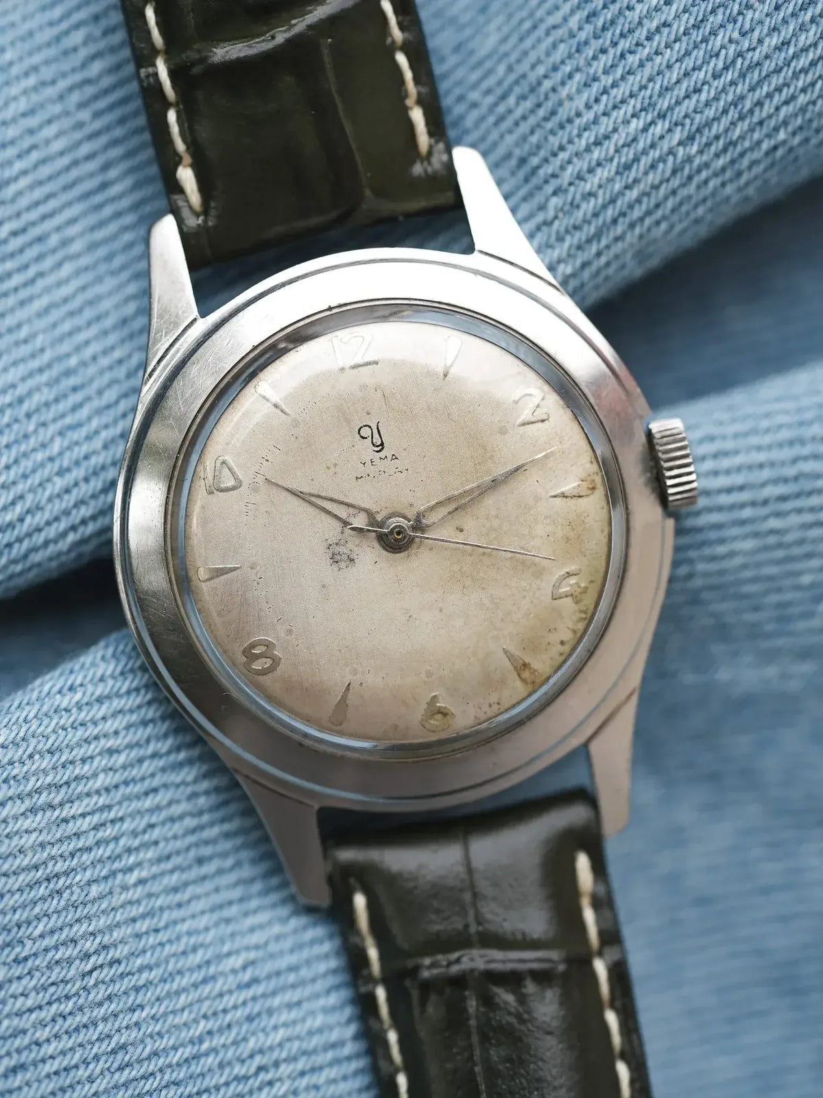 Vintage Yema wristwatch with aged silver dial, black leather strap, on denim background