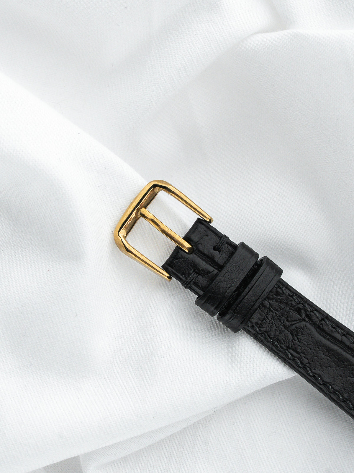 Boucle de montre dorée sur bracelet en cuir noir grainé, fond blanc satiné