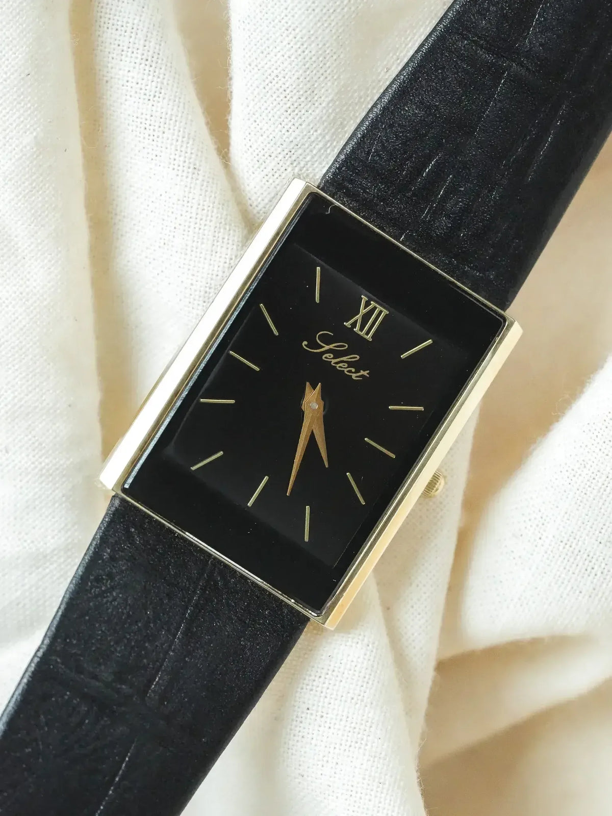 Select vintage watch with black dial, gold case, and black leather strap on fabric
