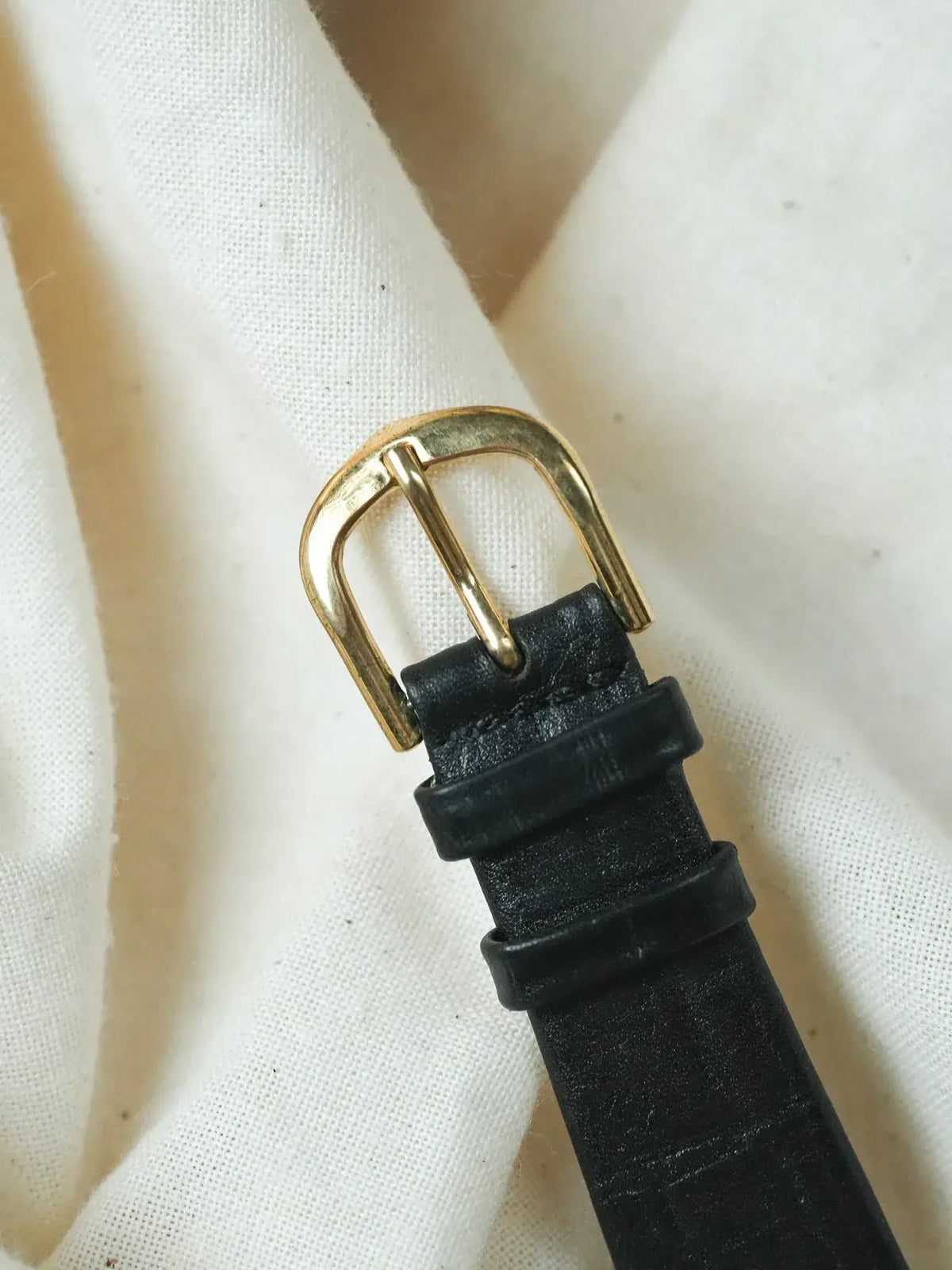 Black leather watch strap with gold buckle on white fabric background