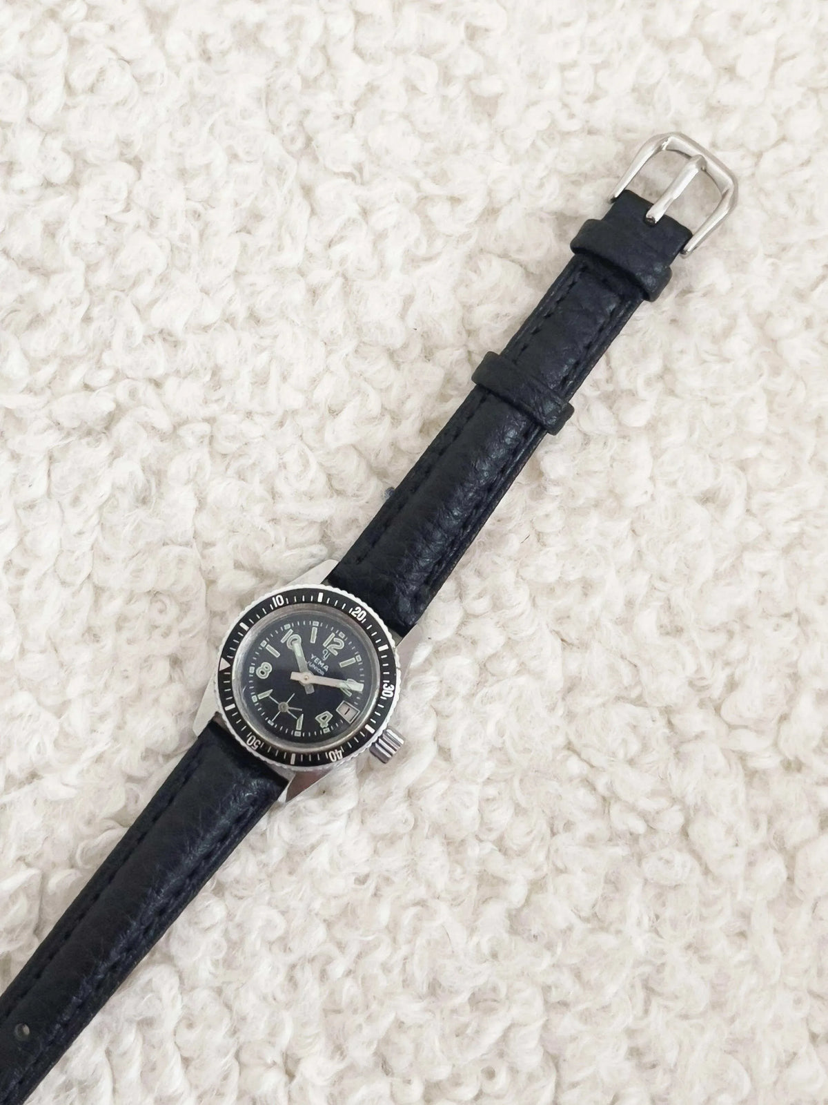 Vintage Yema Junior watch with black dial, silver case, and black leather strap on textured white background